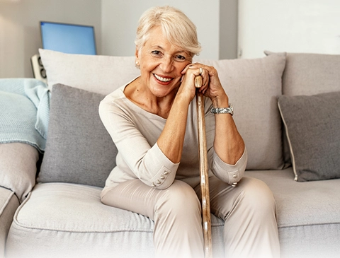 Eine Senioren sitzt gemütlich zu Hause mit einem Gehstock in der Hand